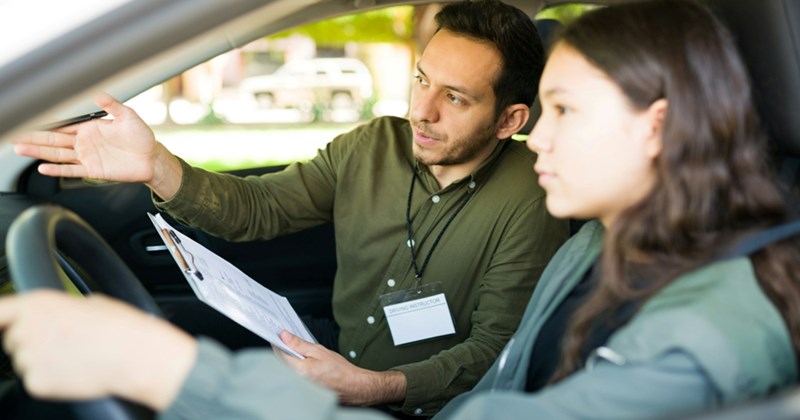 A male driving instructor holding a clipboard instructs a teenage female learner driver in a vehicle