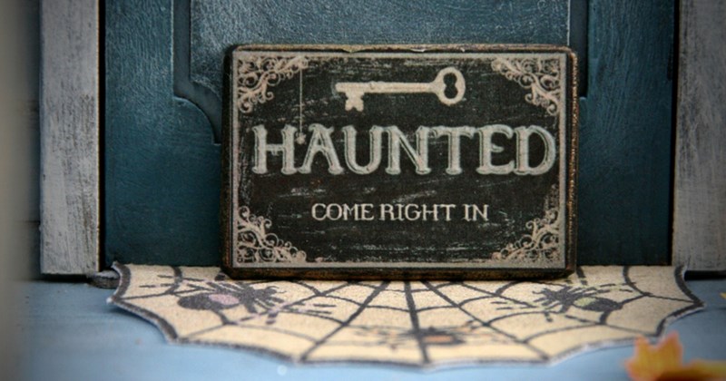 A doormat with a cobweb and spider pattern on it in front of a door with a sign at the bottom that says 'haunted, come right in'