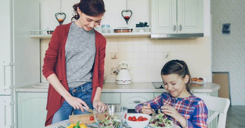 A mom stands at a kitchen table cutting tomatoes while her daughter sits next to her, eating salad