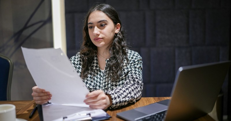 An employee looks at a piece of paper while sitting at a table with her laptop beside her