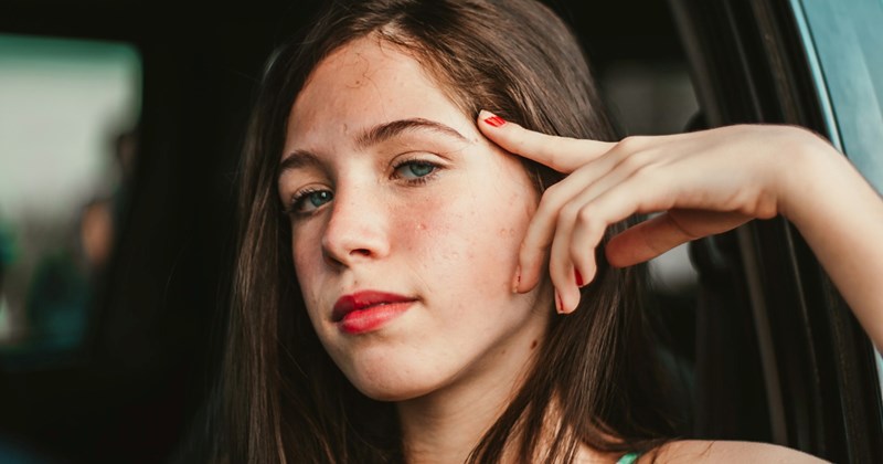 A girl rests her arm on an open car window, touching her face with her hand