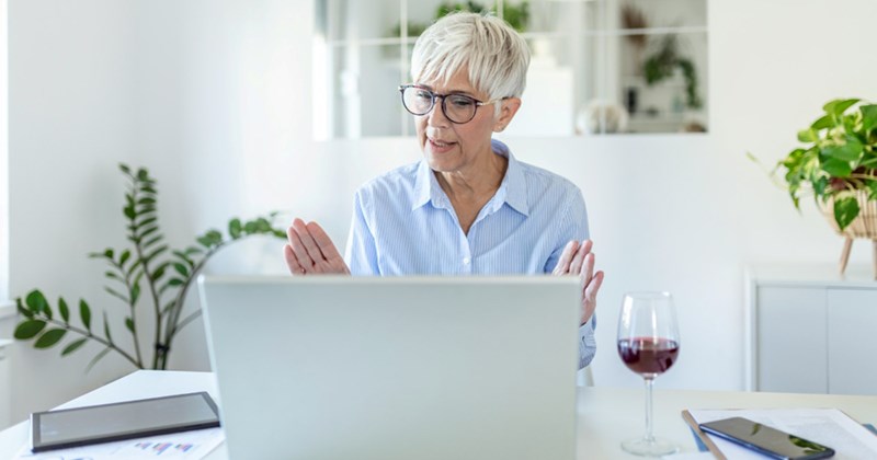 An older employee takes a meeting on her laptop at home with a glass of wine by her side
