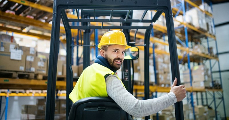 A man wearing a hard hat looks behind him and smiles while sitting in a forklift in a warehouse