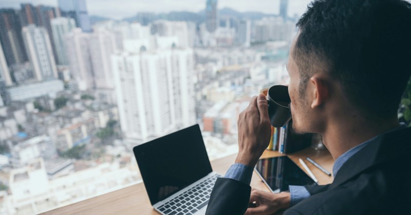 A businessman, seen from behind, drinks from a mug and works at his laptop with a view of the city skyline