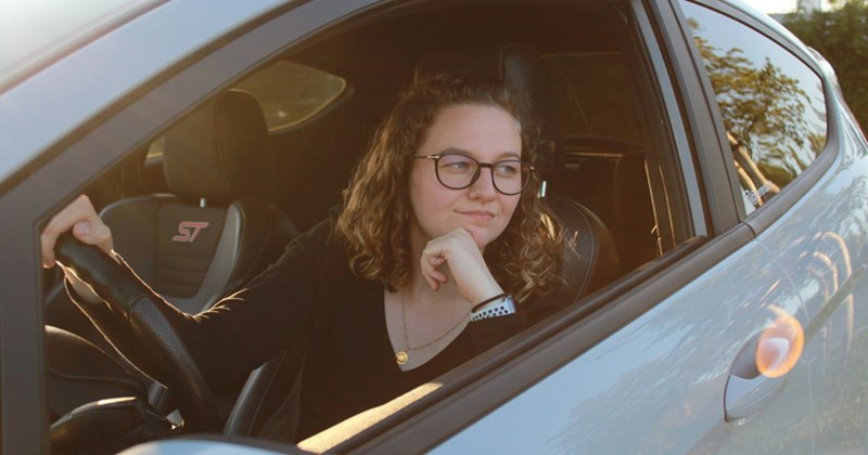 A woman wearing glasses sits in the driver's seat of a car with the window open