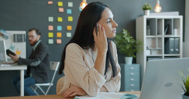 A female employee touches her face as she sits behind a laptop and an open planner in an open-plan office 