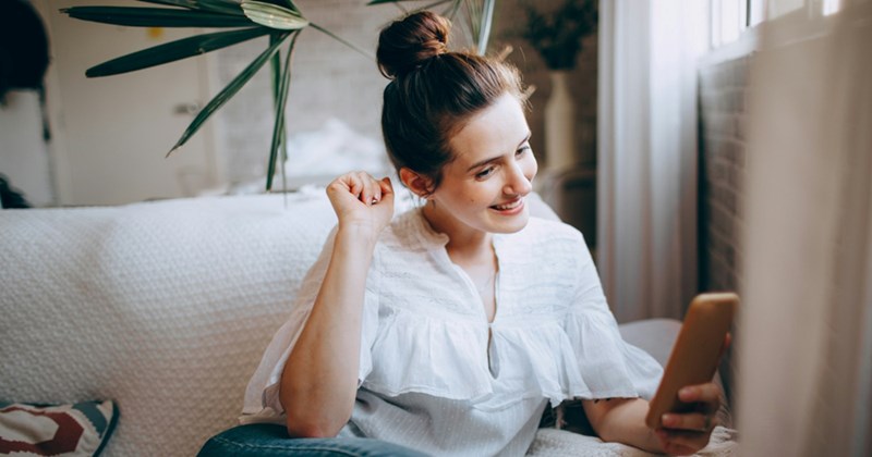 A woman sitting on a couch smiles and holds up her hand while looking at her phone