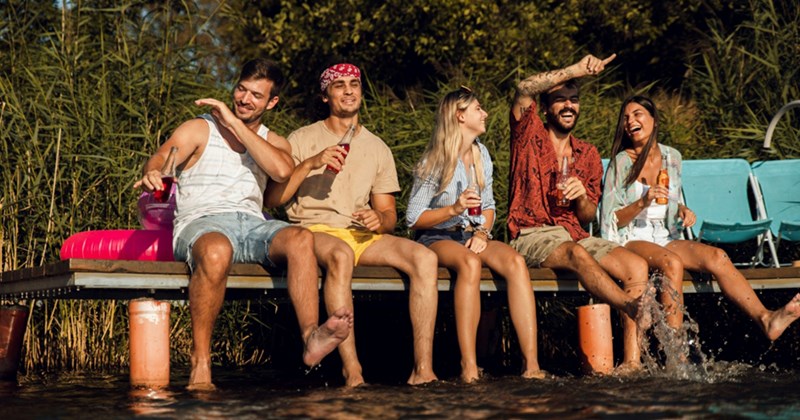 A representation of five male and female friends sitting on the pier of a lake, smiling and holding drinks