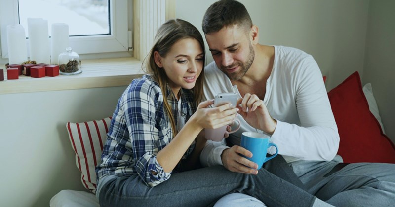 Models representing a male and female couple sitting on a couch together, the man holding a mug as he looks at something on the phone the woman is holding