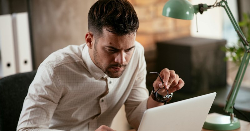 A representation of a male employee staring at his laptop, confused, holding a pair of glasses in one hand