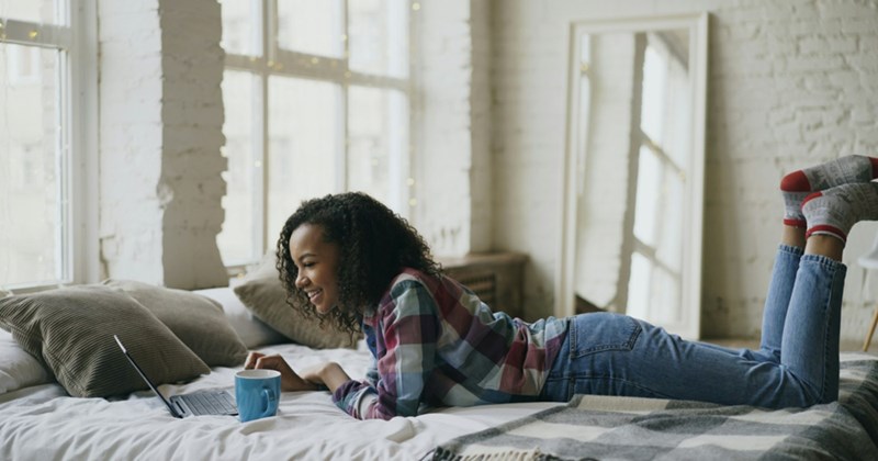 A representation of a young woman lying on top of a bed, using a laptop with a mug beside her