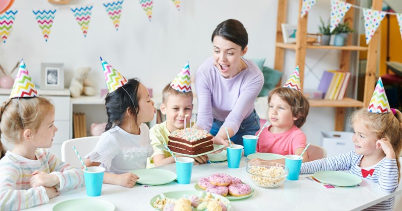 A representation of a mom holding a birthday cake out to her daughter, who is sitting at a table with other children wearing party hats