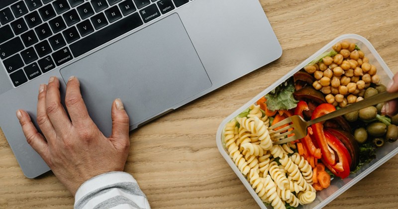A representation of a person's hand resting on an open laptop, with a Tupperware containing pasta salad beside them