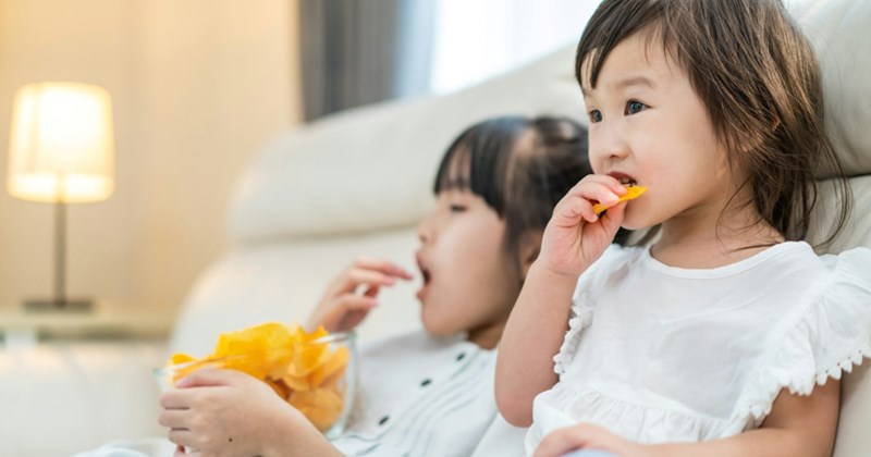 A representation of two young girls eating chips from a bowl while sitting on a couch