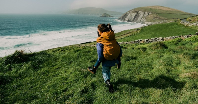 A representation of a man piggybacking a girl down a grassy hill towards the sea with a cliff in the background