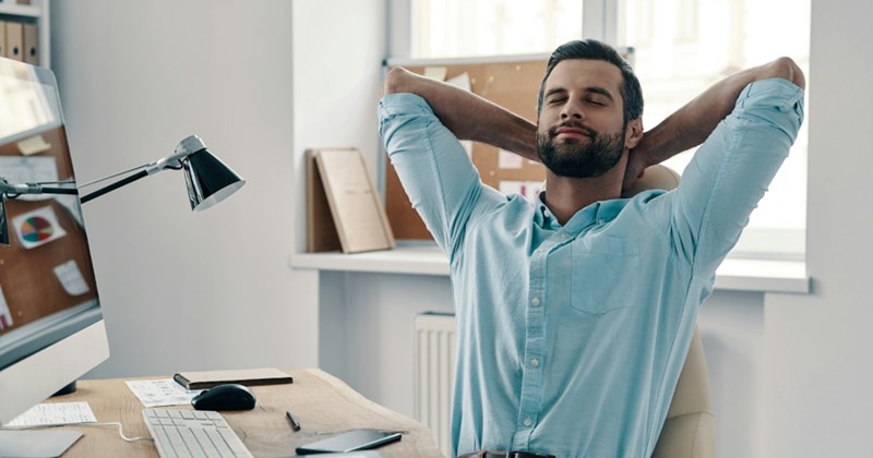 A representation of an employee sitting at a computer desk, closing his eyes and clasping his hands behind his head