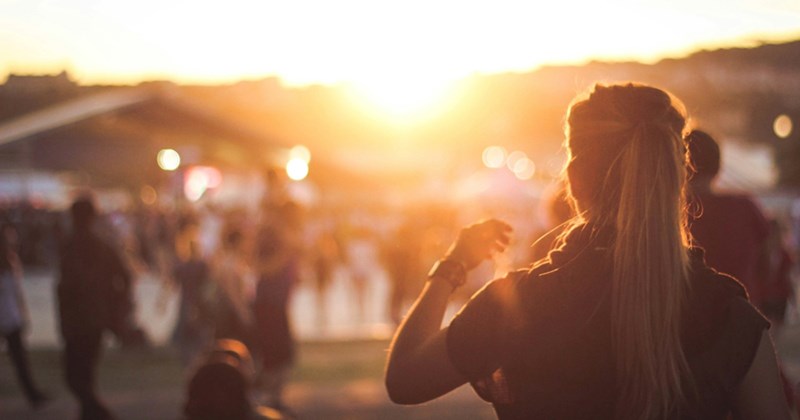 A representation of a woman with her back turned, looking into the crowd gathering for an outdoor concert and the sunset