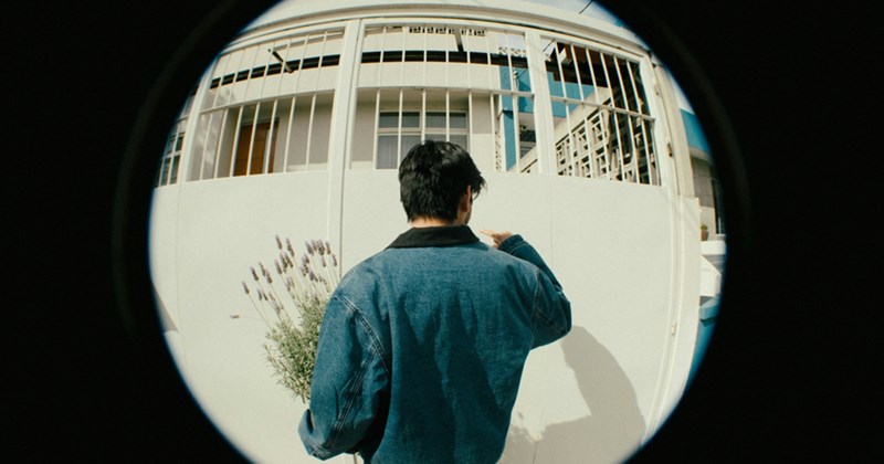 The back of a man holding a lavender plant and knocking on a front door, as seen through a fisheye lens