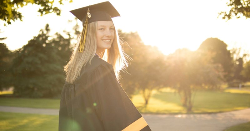 A model representing a graduating high school student, looking over her shoulder while wearing a cap and gown outside