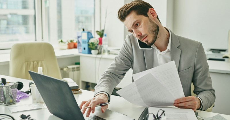 A representation of an employee holding papers, looking at something on his laptop, and cradling a cell phone to his ear