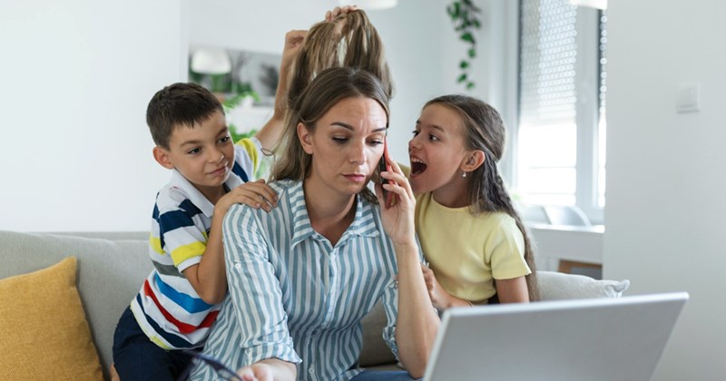 A representation of a mom on her cellphone sits on a couch, working on her laptop as two kids try to get her attention on either side of her