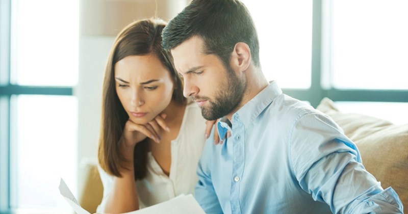 A male and female couple sit on a couch together, looking at documents