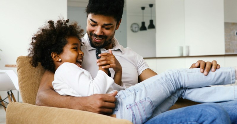 A young girl laughs while sitting on her father's lap on a couch