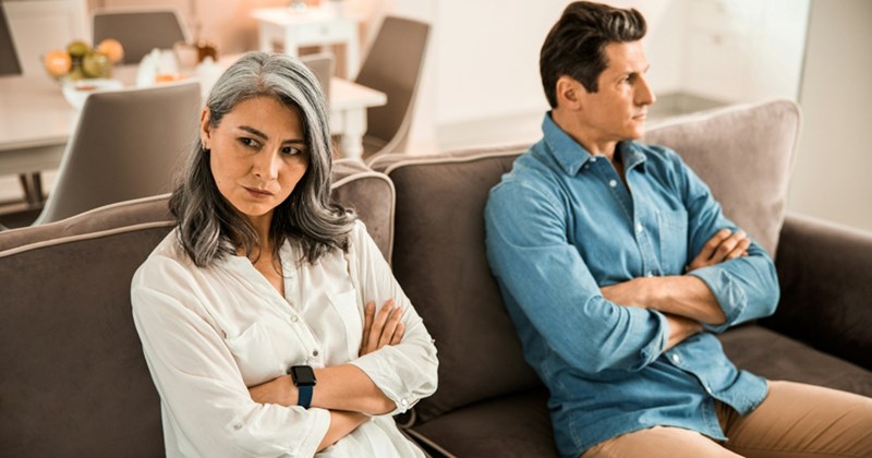 A man and a woman sit at opposite ends of a couch, looking away from one another with their arms crossed