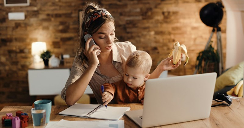 An employee works from home with a baby on her lap, talking on the phone, and holding a half-eaten banana