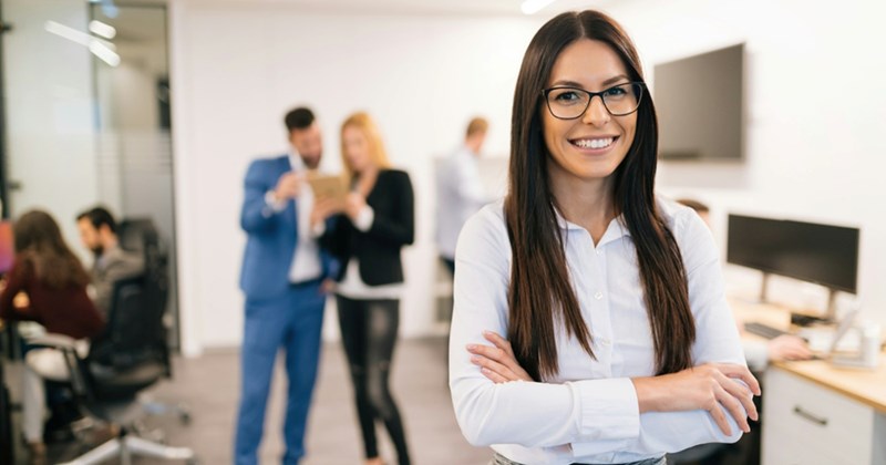 An employee crosses her arms and smiles while standing in an office with colleagues having a discussion in the background