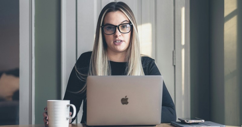 A representation of a female manager sitting behind a MacBook while wearing glasses and holding a mug beside her 