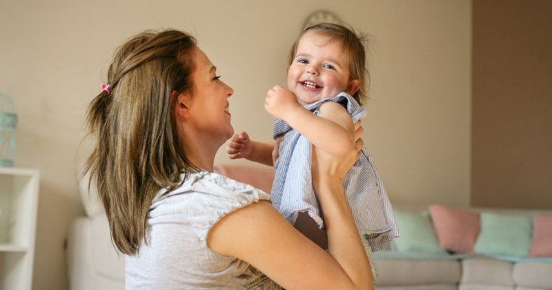 A toddler smiles as a woman lifts her up in her room