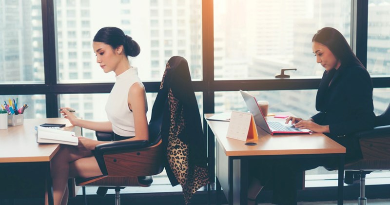 Two female employees work at desks in an office, one in front of the other