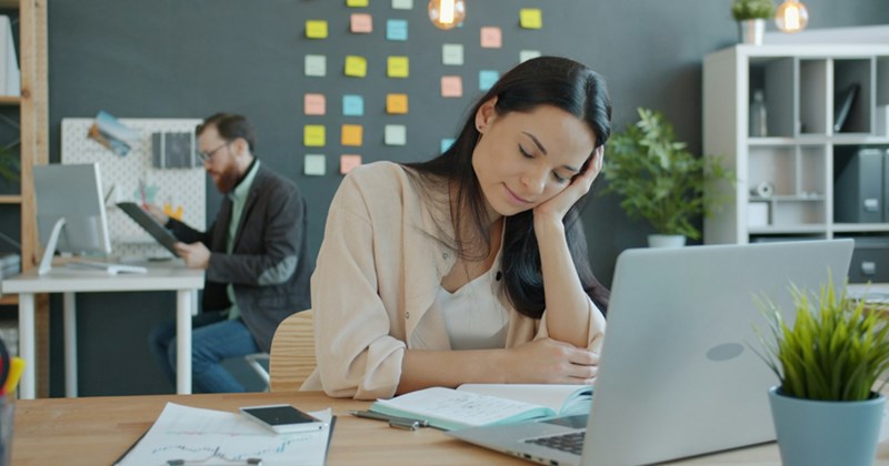 A representation of an employee closing her eyes and resting her head in her hand while sitting behind a laptop, with a colleague working in the background