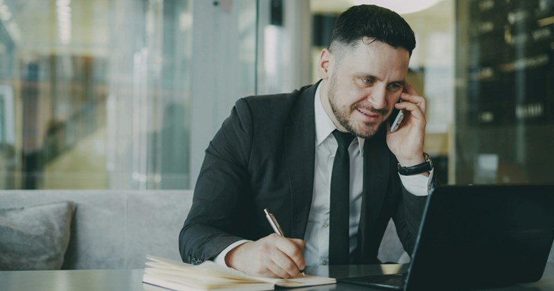 A representation of a man in a suit sitting behind a laptop, talking on a cell phone, and making notes in a notebook