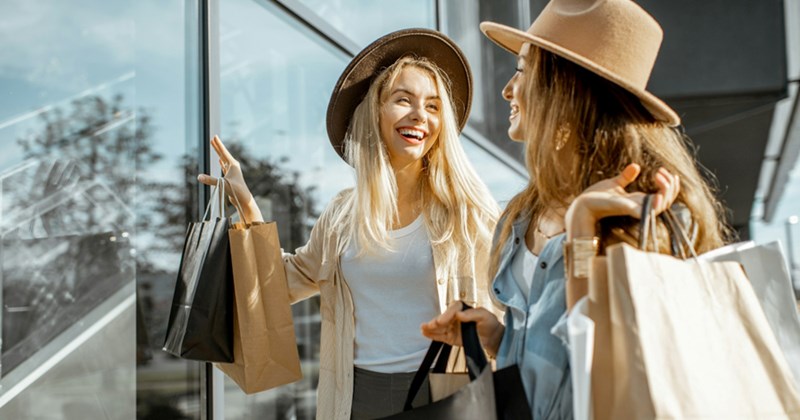 A representation of two female friends wearing wide-brimmed hats and holding shopping bags, talking to each other next to a glass-fronted building