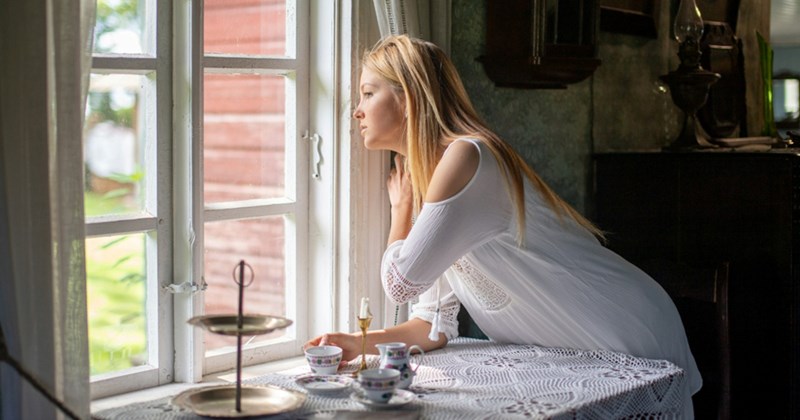 A representation of a woman peering out of a window over a table with a tea set on it