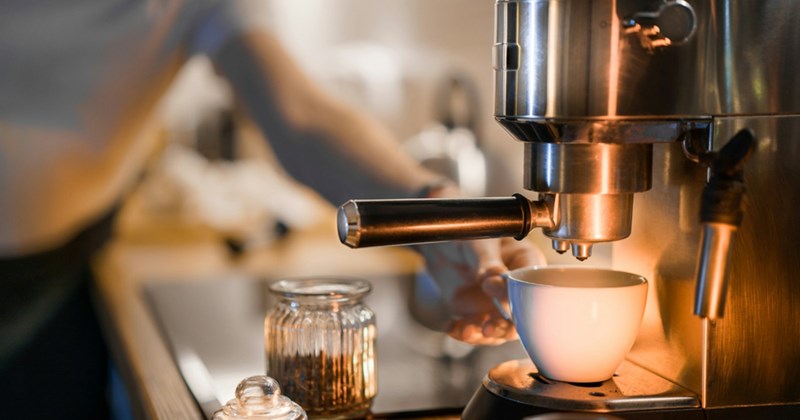 A coffee machine on a counter with a person reaching towards it in the background