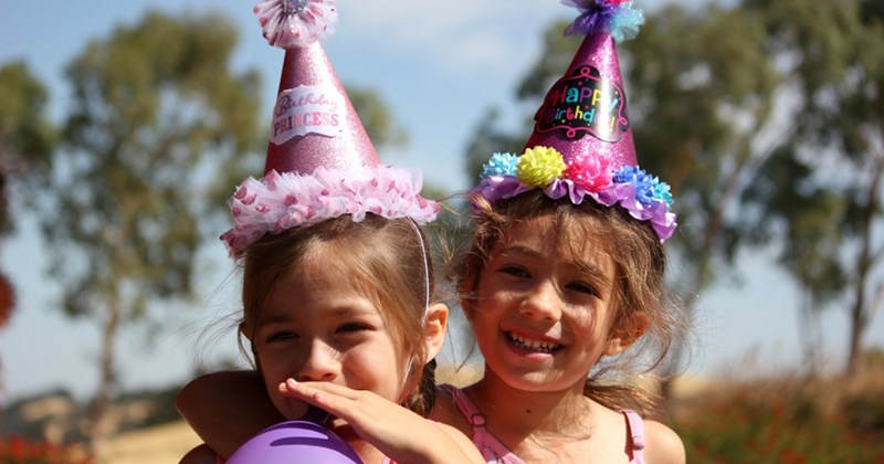 Close-up of two young girls standing side by side, wearing party hats, with one blowing a balloon 