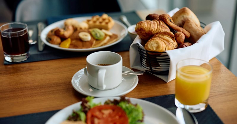 A close-up of a table with a hotel breakfast, including a basket of pastries, cups of coffee and juice, and plates with pancakes, waffles, and salad on them