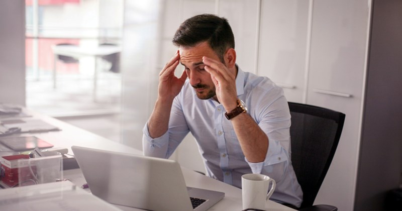 An exasperated employee rubs his temples while sitting at his work desk