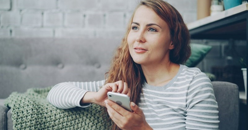 A woman looks into the distance while sitting on her couch and using her phone