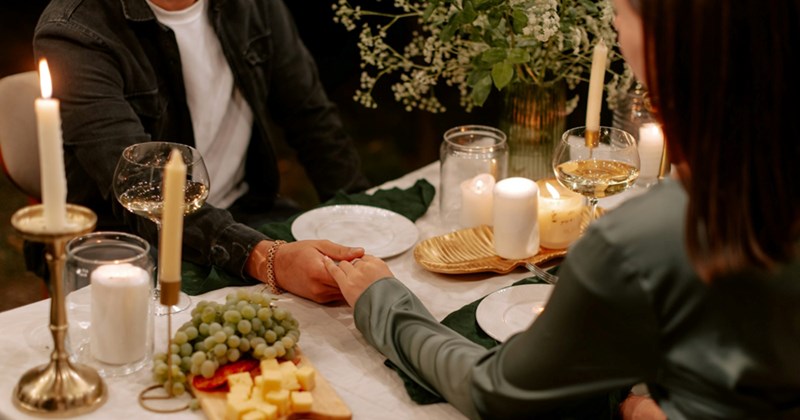 A man and a woman hold hands across a candlelit dinner table with a cheeseboard and wine glasses