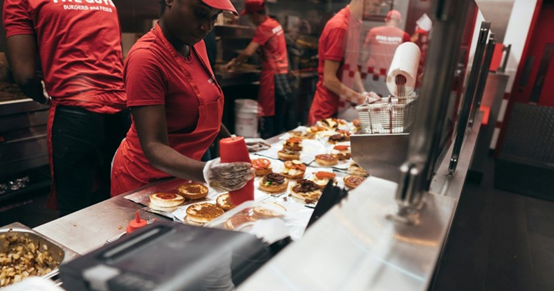 A representation of a fast food worker with several open burgers in front of her, squeezing sauce onto a bun
