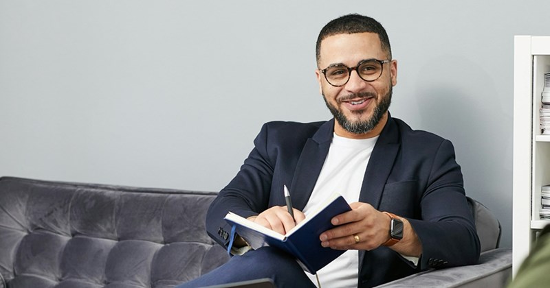 A professional man smiles as he sits on a black leather couch, writing in a notebook