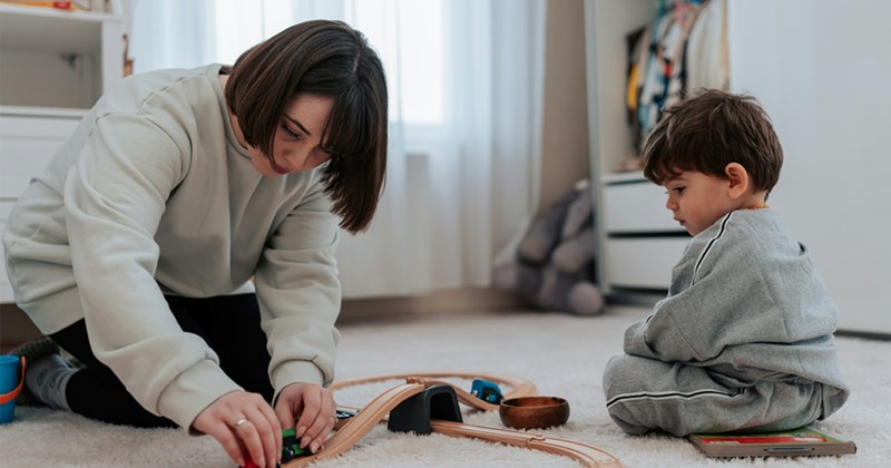 A representation of a woman and a toddler playing with a wooden train set on the floor