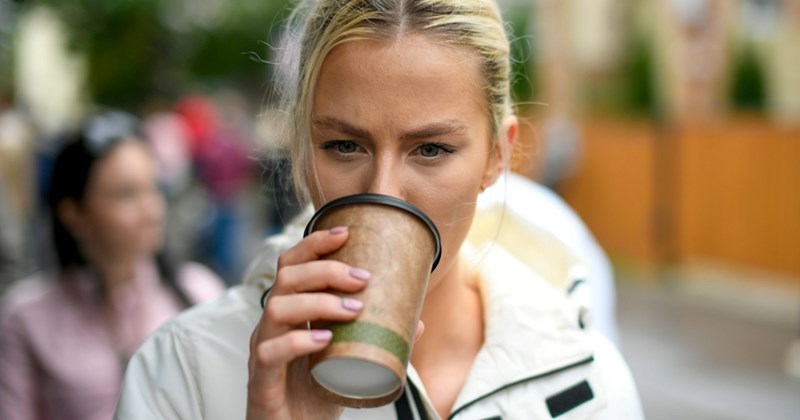 A model representing a blond woman drinks from a disposable coffee cup while standing in the street