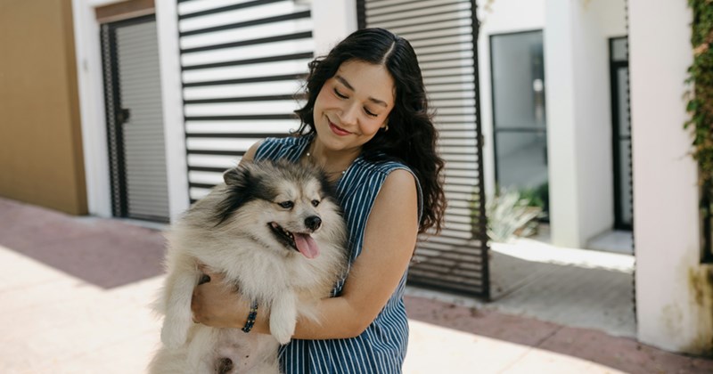 A woman smiles while looking down at the small dog she is holding while standing outside a building