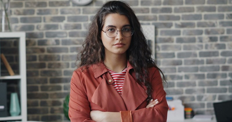 A representation of a female employee wearing smart casual, standing in an office and frowning with her arms crossed