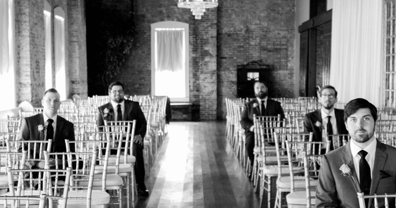 Grayscale photo of several male wedding guests sitting in rows of chairs arranged for a wedding ceremony. 